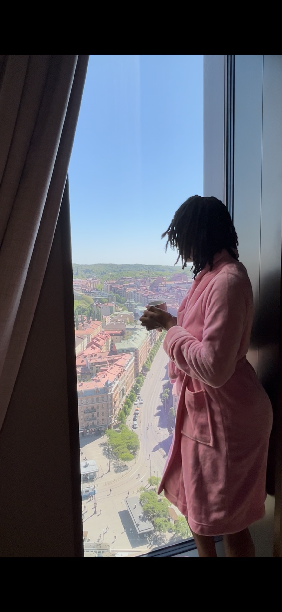 Stefanee Richardson stands in a pink robe by a high-rise window, holding a cup of coffee and looking out over the city of Gothenburg, Sweden.